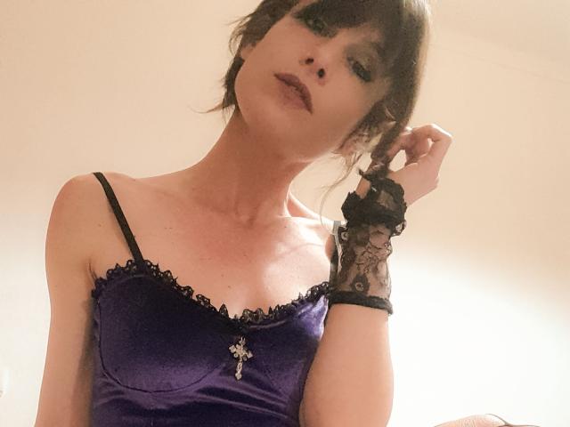 Aiyana with dark hair, purple satin top, black lace wrist, and cross necklace, looking down, touching her hair, showing her tits. Her eyes are half-closed.