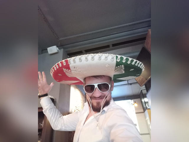 PasitiF trägt einen riesigen mexikanischen Sombrero, weiße Sonnenbrillen und ein weißes Hemd, winkt lächelnd, hat einen Bart und trägt weiße Kopfhörer..