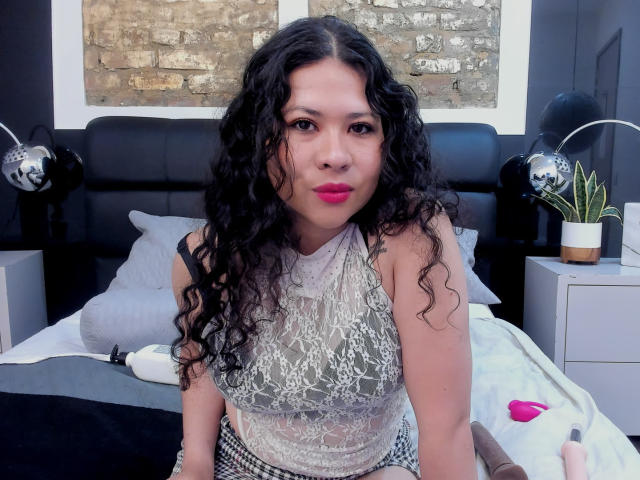 LourrieAnn mit lockigem, schwarzem Haar, die ein Spitzenoberteil trägt, sitzt auf einem Bett mit einem Dildo und einer Rose, trägt rosafarbenen Lippenstift, im Hintergrund ist ein schwarzer Kopfteil und eine Zimmerpflanze zu sehen..