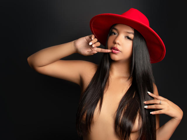 TayraBankss, senza top e con lunghi capelli neri, indossa un cappello rosso e punta le dita sulle sue labbra rosa, su uno sfondo scuro. Il suo seno è visibile, non indossa altri vestiti..
