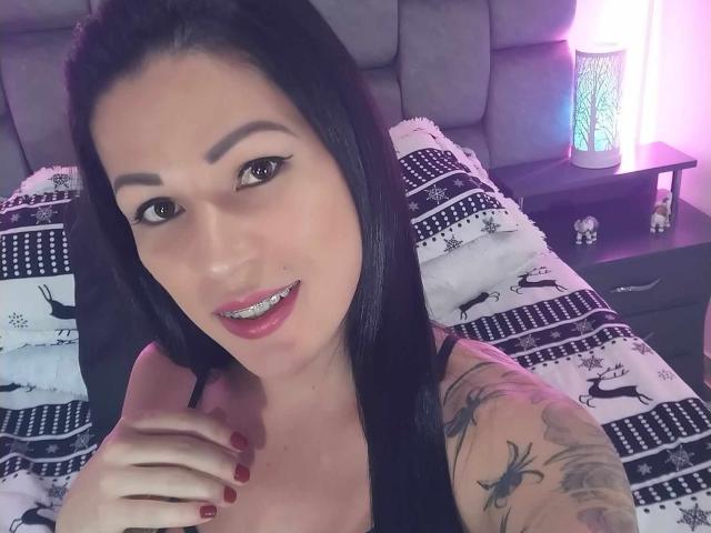 SexyCaandydoll with black hair, brown eyes, red lipstick, and tattoos on her right arm, smiling at the camera, holding her chest with red-nail painted fingers.