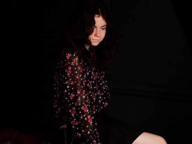 TinaRedly with curly brown hair, wearing a black floral blouse, sitting on a red surface, showing her ass, black skirt, and a subtle, confident smile in dark lighting.