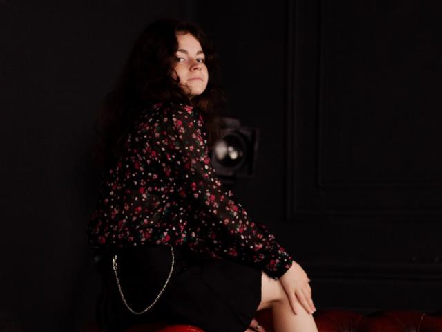 TinaRedly, com cabelos escuros e cacheados, vestindo uma blusa preta com estampas florais e uma saia preta, sentada em um sofá vermelho, olhando por cima do ombro, com a mão na coxa, e segurando uma bolsa de corrente..
