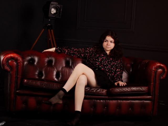 TinaRedly with curly brown hair, wearing a black floral blouse and black shorts, sits on a red leather couch, legs crossed, black socks, looking at the camera.