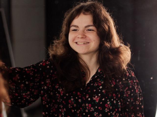TinaRedly with wavy brown hair, smiling, wearing a black floral dress, has fair skin and is holding an arm out. She's standing in a dimly lit area.