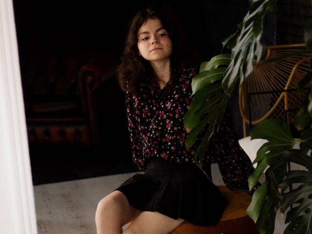 Tina, mit lockigem, braunem Haar, trägt eine schwarze Bluse mit Blumenmuster und einen schwarzen Rock, sitzt auf einem Holztisch, neben einer grünen Pflanze, mit ernstem Gesichtsausdruck..