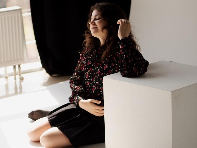 TinaRedly with curly brown hair, wearing a floral black shirt and black skirt, sitting on white cube, smiling, right hand in hair, left hand on cube, sunlight.