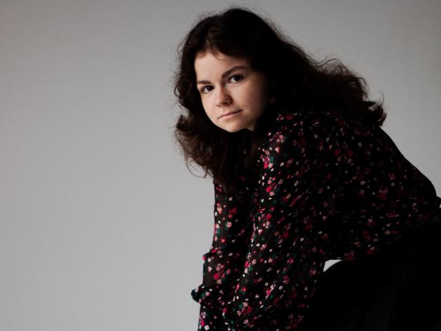 TinaRedly with curly dark hair, wearing a black floral shirt, looking sideways with a slight smile, her ass barely visible in the lower right.