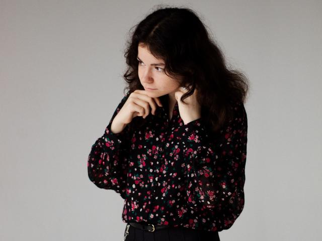 TinaRedly with wavy dark hair, wearing a black floral blouse, looking to the side, holding her chin with one hand, showing her bare ass with black pants.