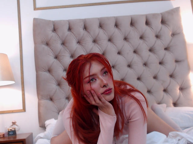 MikyValkova, com cabelo ruivo, suéter rosa e piercing no nariz, deitada na cama, com a mão no rosto, olhando para a câmera, com a parte de trás do corpo levemente visível, cabeceira com textura, iluminação suave, expressão relaxada..