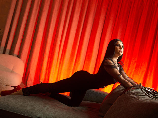 MelissaGlow in black bodysuit, on gray couch, red high heels, leaning forward, red curtains, tattoos on arm, side profile, looking to right, sexy, seductive, glowing.