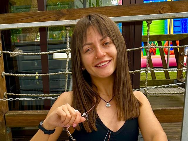 LikaLuis has brown straight hair, smiling with pink lips, holding glasses in her right hand, wearing a black top, necklace with a silver pendant, and a black wristband.