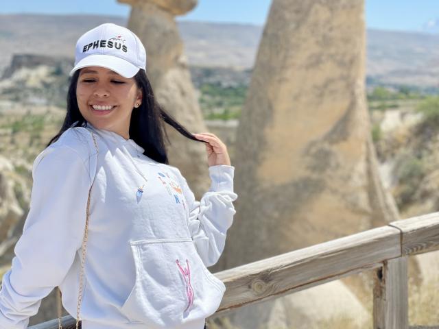 MeganWisse with long black hair, white cap, and white hoodie, smiling, holding hair, standing by wooden railing, rock formations in background, sunny day.