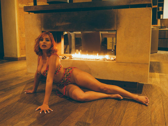GalaMalkova with pink hair, floral bikini, large tits, and a curvy ass, laying on wooden floor in front of a lit fireplace, looking at the camera.