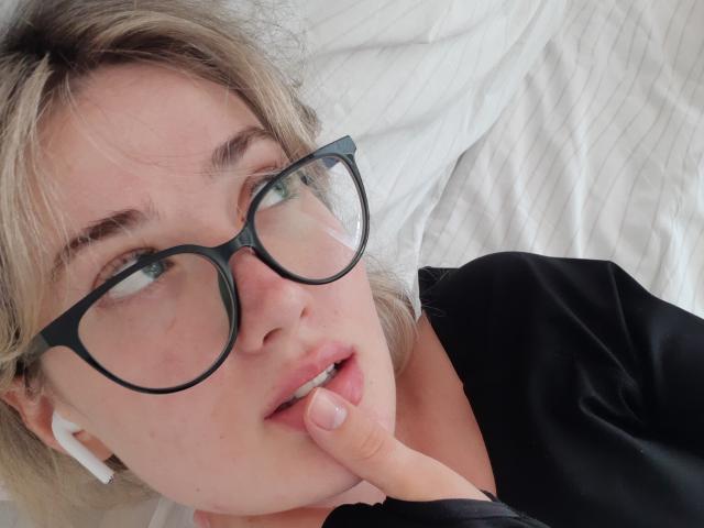 Cindy, avec ses cheveux blonds, ses lunettes noires et son t-shirt noir, mordille son doigt, porte des écouteurs blancs, a une peau claire et des yeux verts, et est allongée sur un lit blanc..