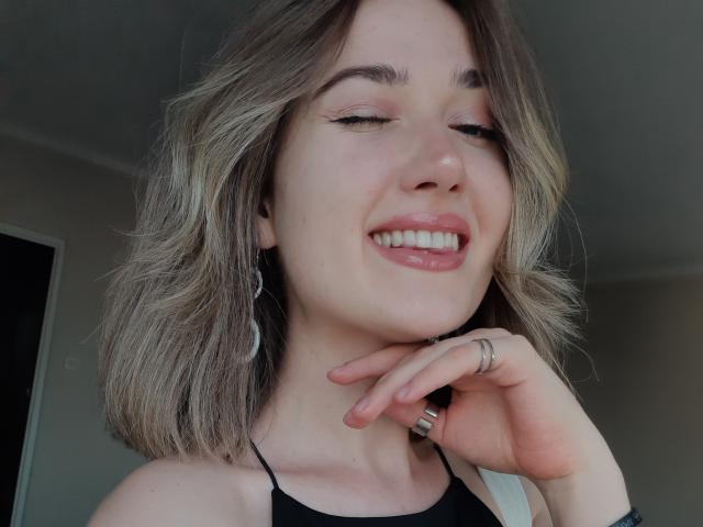 Cindy, avec des cheveux blonds ondulés de longueur moyenne, souriant les yeux fermés, portant un haut noir à encolure dégagée, une bague à un doigt, des boucles d'oreilles argentées et un maquillage discret, dans un selfie décontracté..