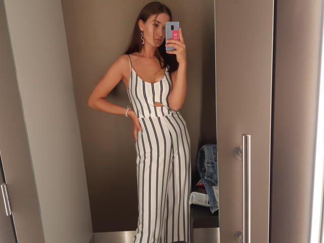 CindyFantasy, avec de longs cheveux bruns, portant un ensemble pantalon-chemise blanc et noir à rayures, découpé, taille haute, tenant un téléphone, montrant sa petite poitrine, et une veste en jean dans le miroir..