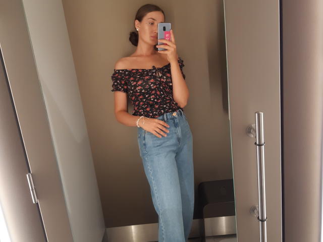 CindyFantasy, avec de petites seins, portant un haut floral à une épaule, un jean bleu taille haute et un bracelet, prend un selfie devant un miroir dans une pièce..