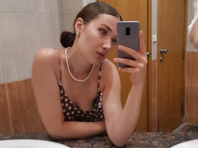 CindyFantasy trägt ein braunes Oberteil mit Punkten, eine Perlenkette und Ohrringe und macht ein Selfie vor einem Spiegel, wobei ihre Brüste zu sehen sind. Sie befindet sich in einem Badezimmer mit einer Holztür..