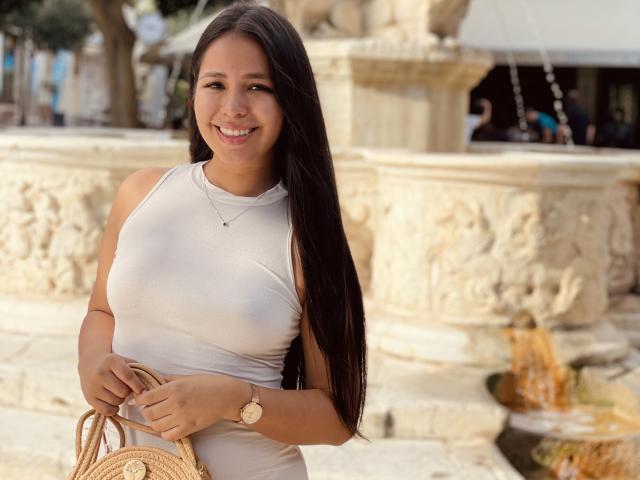 Megan Wisse, con lunghi capelli neri, sorridente, indossa una canotta bianca aderente che mette in mostra i suoi piccoli seni, tiene una borsa di vimini, un orologio d'oro e una collana, ed è in piedi accanto a una fontana decorata..