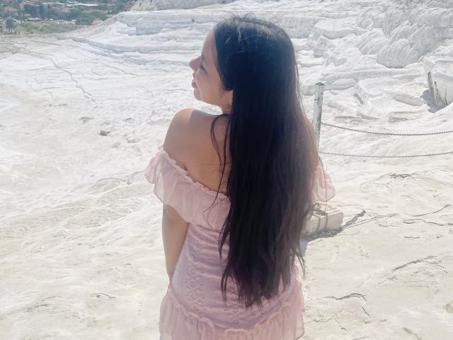 MeganWisse with long black hair, wearing a pink off-shoulder dress, standing in a white, rocky, outdoor area, looking to the side, showing her back and a small ass.