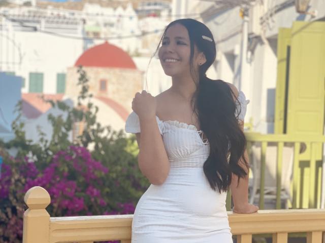 Megan Wisse, com longos cabelos pretos, vestindo um vestido branco com um ombro descoberto, sorrindo, segurando o cabelo, em pé perto de um corrimão de madeira, com uma porta amarela ao fundo e flores rosa..