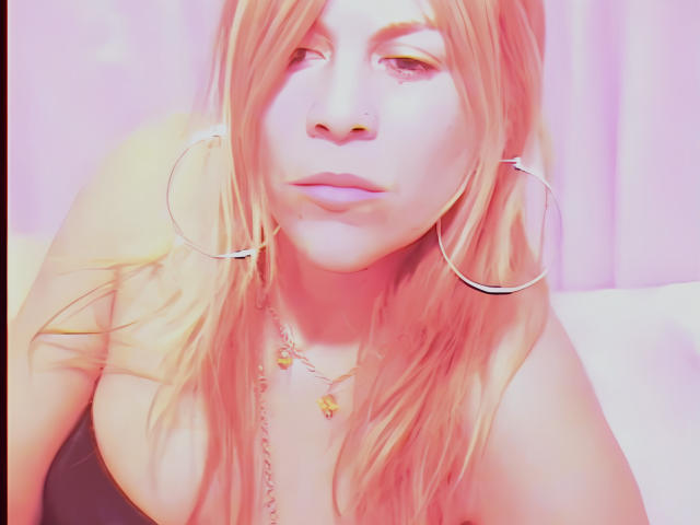 Maytena with pinkish-orange hair, large hoop earrings, and a necklace, showing off her tits and a nose ring, with a slightly pouty face and soft pink lighting.