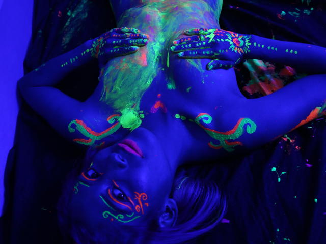 AbyColeman's blue-tinted body covered in neon green and orange paint, lying on black cloth, hands on chest, tits painted, face and hands decorated, blue lighting, paint splashes.
