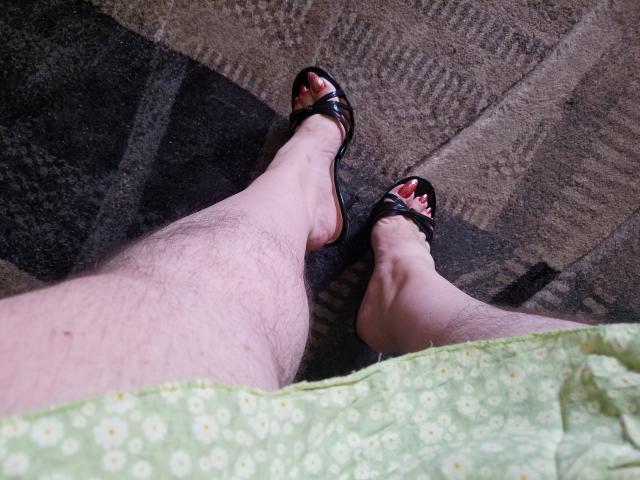 KaryQueen's hairy legs with pale skin in black sandals, toes painted red, wearing a green floral dress, shown from a bottom-up perspective.