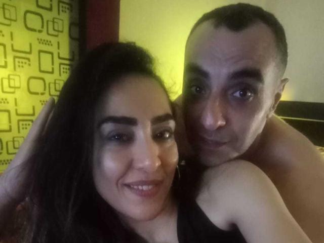 FemlosaZion, com cabelo e maquiagem escura, está sorrindo e usa um piercing no nariz. Ela está abraçando um homem sem camisa e com a cabeça raspada, que parece ligeiramente irritado. O braço dele está ao redor dela..