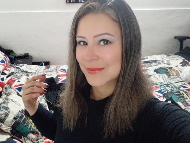 Contelaya with brown hair, black top, red lipstick, and dark nail polish, smiling at the camera, holding her right hand up. Her tits are small, and she's cute.