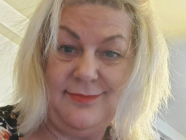 LatexShantie heeft bleke huid, blond, rommelig haar, blauwe ogen en ze glimlacht met rode lippenstift, waarbij een lichte dubbele kin en een kleine neus zichtbaar zijn. Ze draagt een top met bloemenprint..