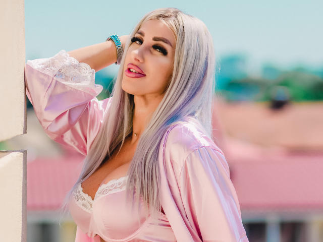TanyaMonroe with long silver hair, pink satin robe, white lace bra, blue bracelet, leaning against a wall, smiling, showing her tits, bright sunlight, pink roof in background.