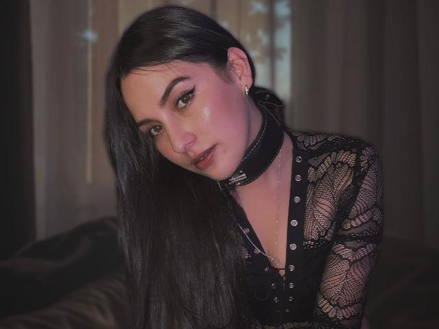 Julianal, con su largo cabello negro, vestida con una blusa de encaje negro y un collar de cuero negro, tiene un rostro ligeramente rosado y mira hacia abajo con una expresión sutil y sensual..