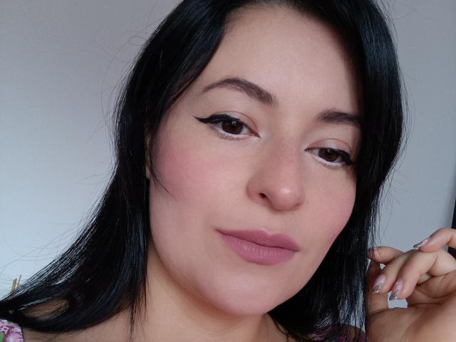 VaaleryStark has black hair, brown eyes, and pink lipstick with winged eyeliner. Her pink cheeks and full lips are prominent. She's touching her hair with manicured nails.
