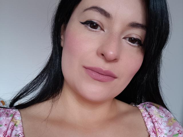 VaaleryStark has black hair, pink lipstick, and winged eyeliner. She's wearing a pink floral dress with puffed shoulders, showing her fair skin and cute, slightly flushed face.
