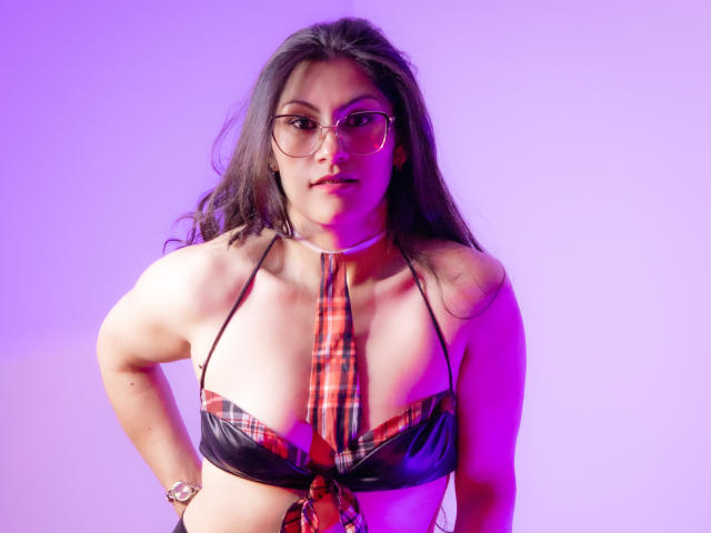 RapZelly avec des lunettes, de longs cheveux bruns, portant une cravate à carreaux et un bikini noir, exposant ses seins, une lumière violette sur son visage, et une montre à son poignet..