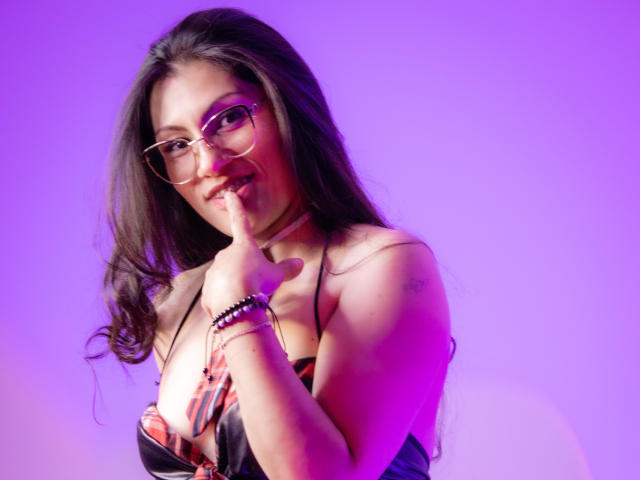 RapZelly, con lunghi capelli scuri, occhiali e un sorriso carino, con un dito sulle labbra, indossa un abito scollato, con luci viola e rosa, e un braccialetto al polso destro..