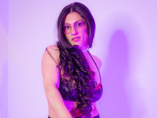 RapZelly, avec ses longs cheveux noirs bouclés, ses lunettes et un éclairage rose-violet, porte un haut noir à carreaux, ressemblant à un soutien-gorge, qui révèle ses seins. Elle regarde la caméra et sourit légèrement..
