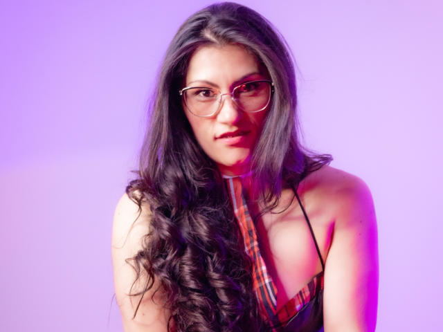 RapZelly, avec de longs cheveux noirs ondulés, portant des lunettes, un haut à carreaux et une cravate rayée, sur un fond violet dégradé, regardant légèrement sur le côté..