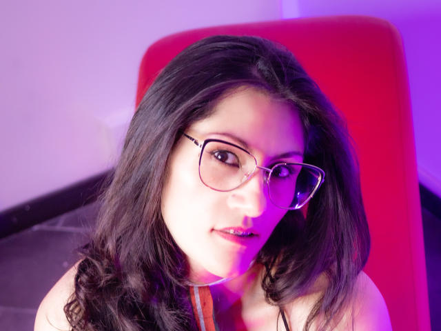 RapZelly with long black hair, glasses, and pinkish-purple light on her face, looking up with a slight smile, her bare shoulders visible, seated on a red chair.
