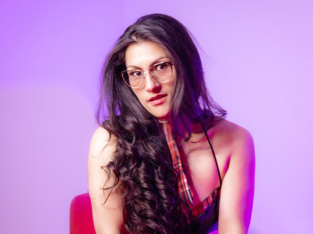 RapZelly, avec ses longs cheveux noirs bouclés, ses lunettes et son haut à carreaux rouges, montrant ses seins, sur un fond illuminé de violet. Elle a un regard mignon et confiant..