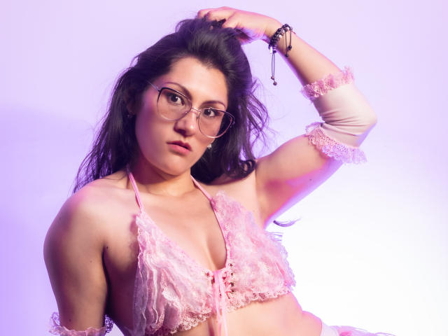 RapZelly com óculos, sutiã de renda rosa e braço levantado, mostrando os seios, usando pulseiras, contra uma luz roxa, com uma expressão séria..