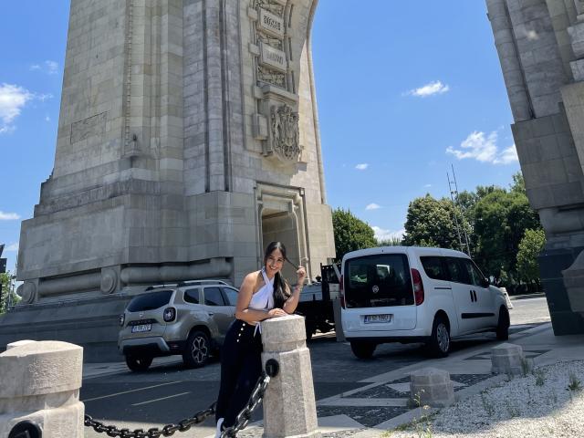 MeganWisse está parada, apoiada em um pilar de pedra, vestindo uma blusa branca sem mangas e calças pretas, com uma corrente na frente, ao lado de carros estacionados e um grande arco de pedra..