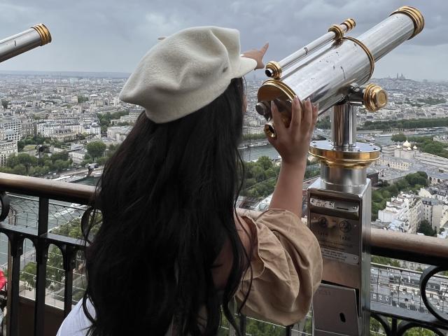 MeganWisse with long black hair, white beret, and beige dress, looking through gold and glass binoculars, standing on a balcony, cityscape in the background, cloudy sky above.