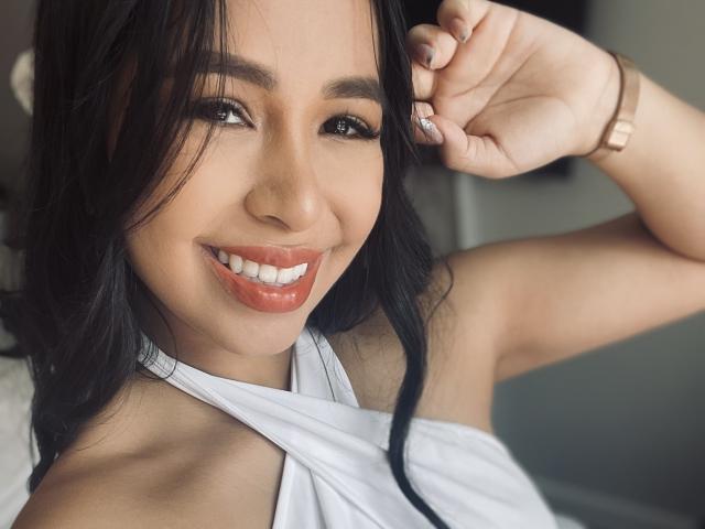 MeganWisse with long black hair, smiling with red lipstick, white halter top, brown wristwatch, and right hand touching hair. She has cute, shiny eyes and small, perky tits.