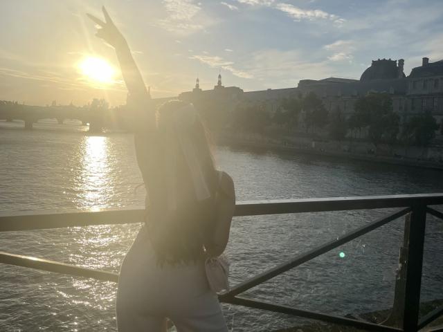 La espalda de MeganWisse está hacia la cámara, con los brazos levantados, el sol brillando a través de ella, vestida con un vestido blanco, de pie junto a un río, con destellos de sol, reflejo del agua, edificios de la ciudad en el fondo..