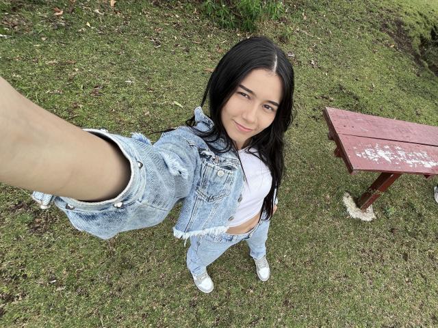 DeisyWest with black hair, white top, and frayed blue jeans, holding out a leg, showing her white sneakers, standing on grass with a red table to the right.