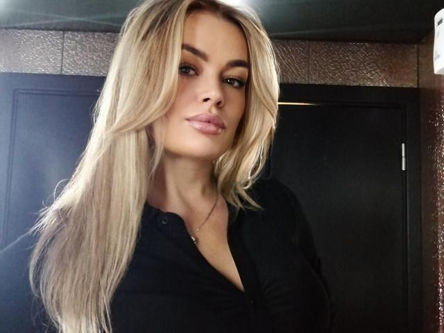 MilenaBiaa, com longos cabelos loiros, vestindo uma camisa preta com botões, tem lábios rosados e um colar de prata, e parece sexy, com os seios levemente visíveis..