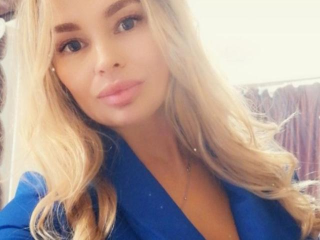 Die blonde MilenaBiaa mit welligen Haaren, blauen Augen und rosafarbenen Lippen, trägt ein blaues Oberteil und hat ein süßes, attraktives Gesicht sowie eine kleine, dezente Halskette..